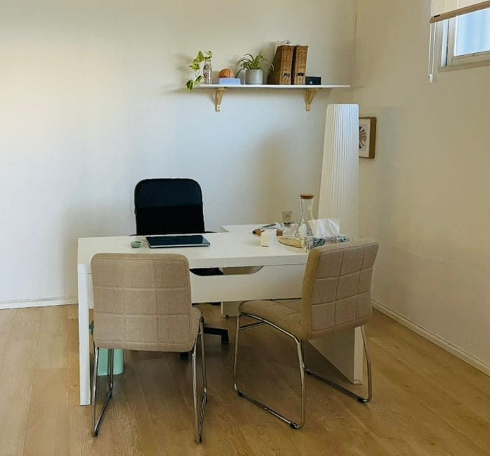 Un escritorio blanco con tres sillas en una oficina minimalista, con una estantería de pared con plantas y carpetas encima.