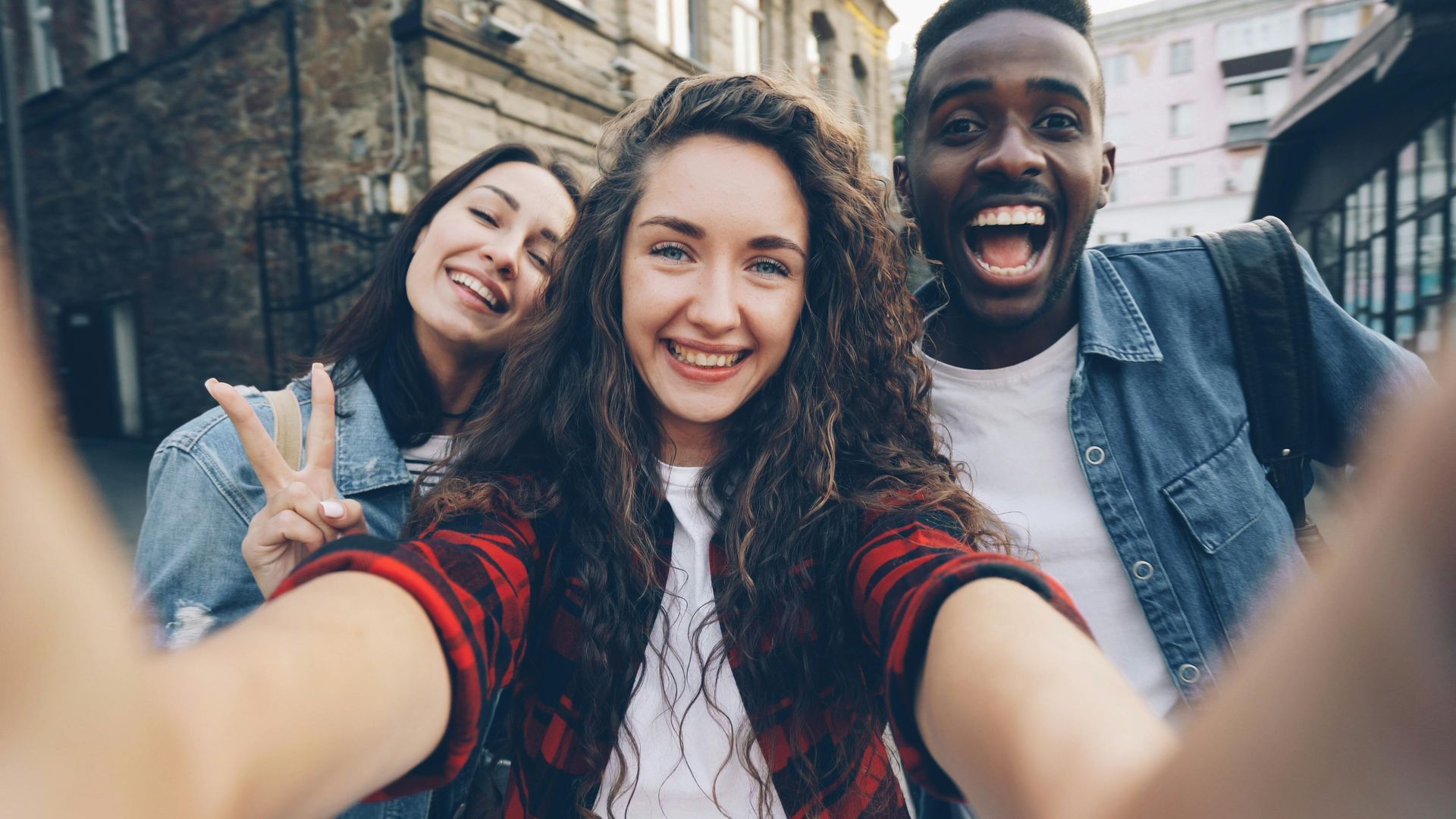 Tres personas sonriendo y tomándose una selfie al aire libre en una calle de la ciudad, una de ellas haciendo el signo de la paz.