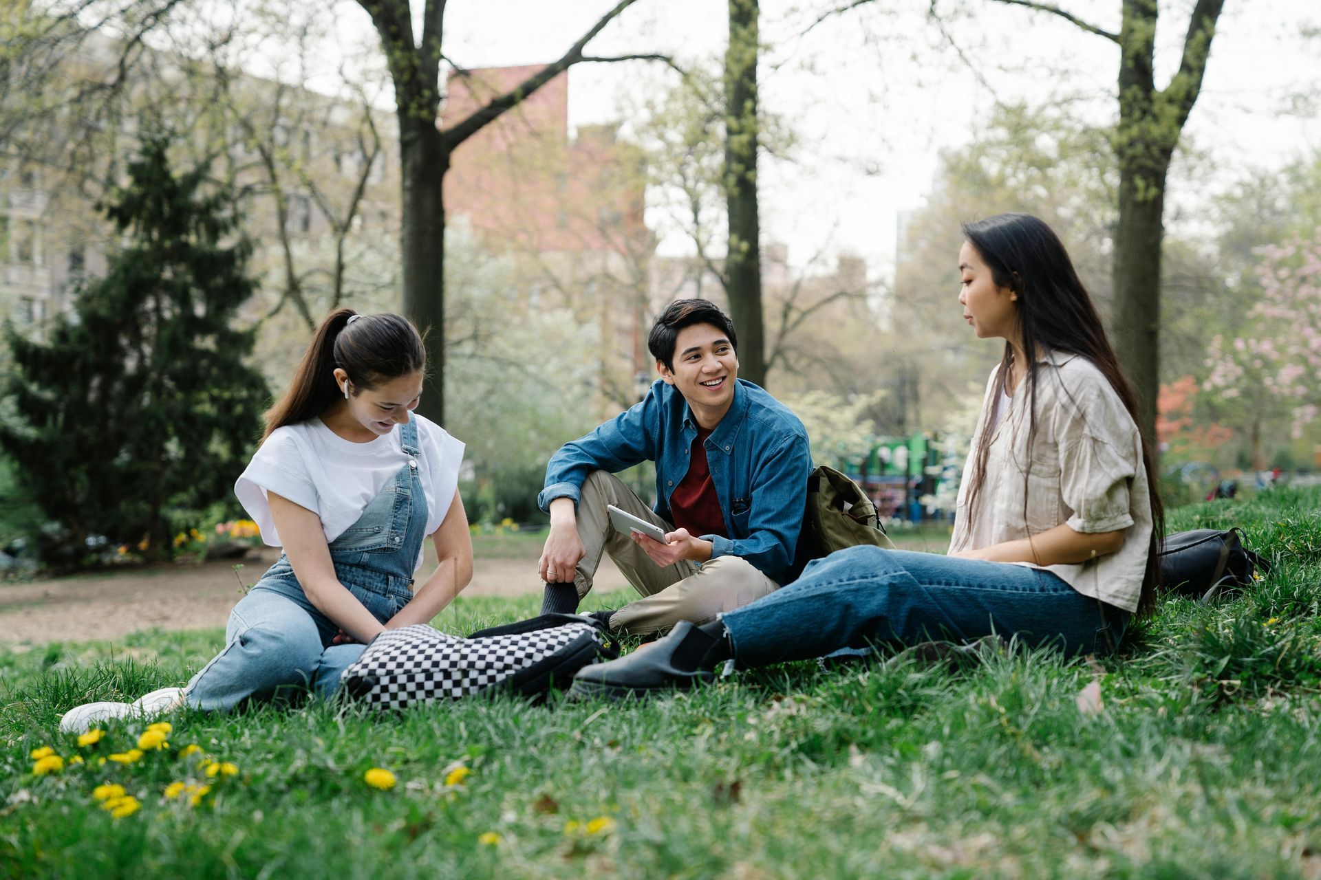 Tres estudiantes están sentados en el césped de un parque, conversando y sosteniendo una tableta, rodeados de árboles.