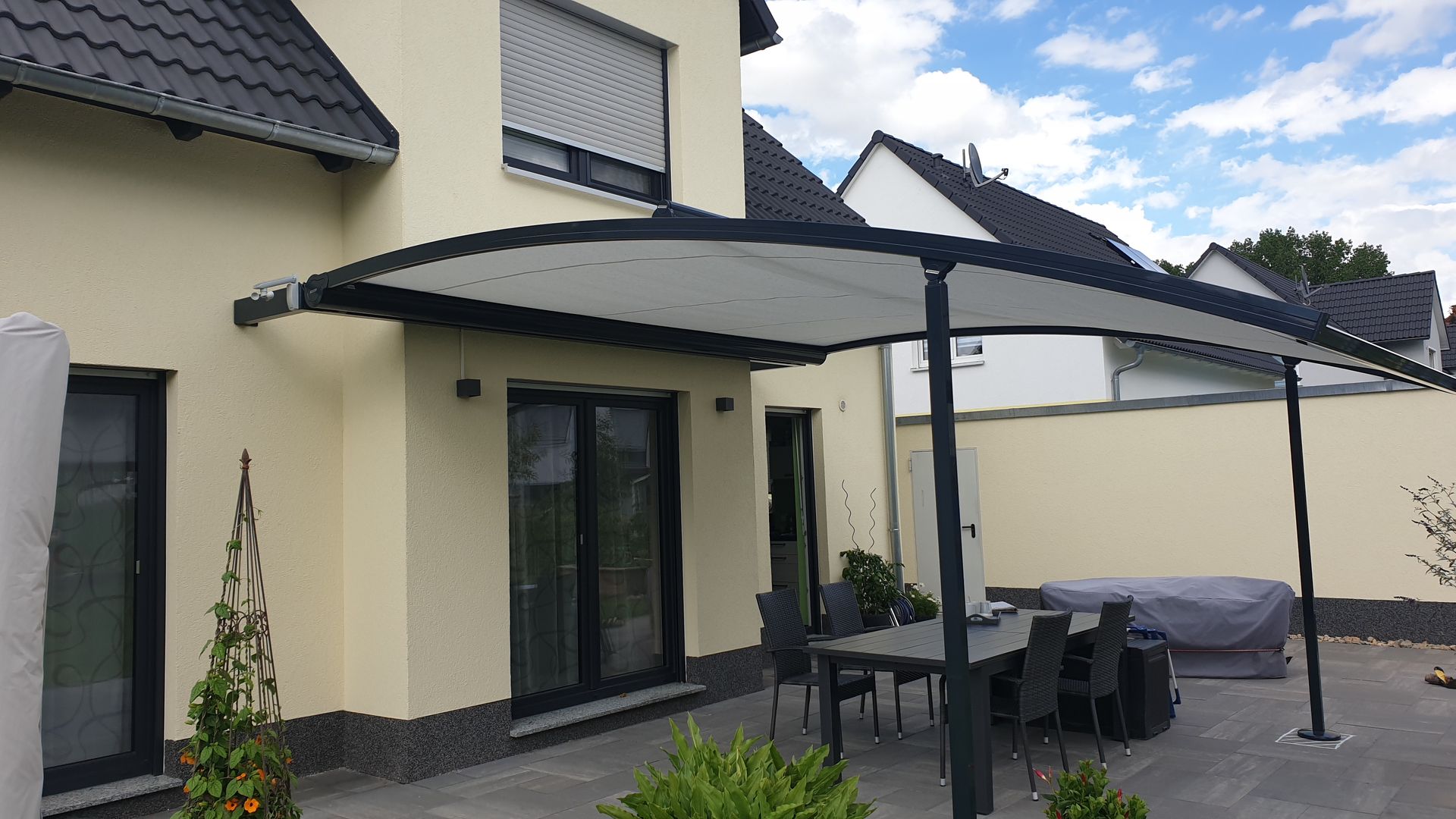 Ein Patio mit einem weißen, bogenförmigen Vordach, der an ein cremefarbenes Haus angrenzt, darunter befinden sich ein Tisch und Stühle.