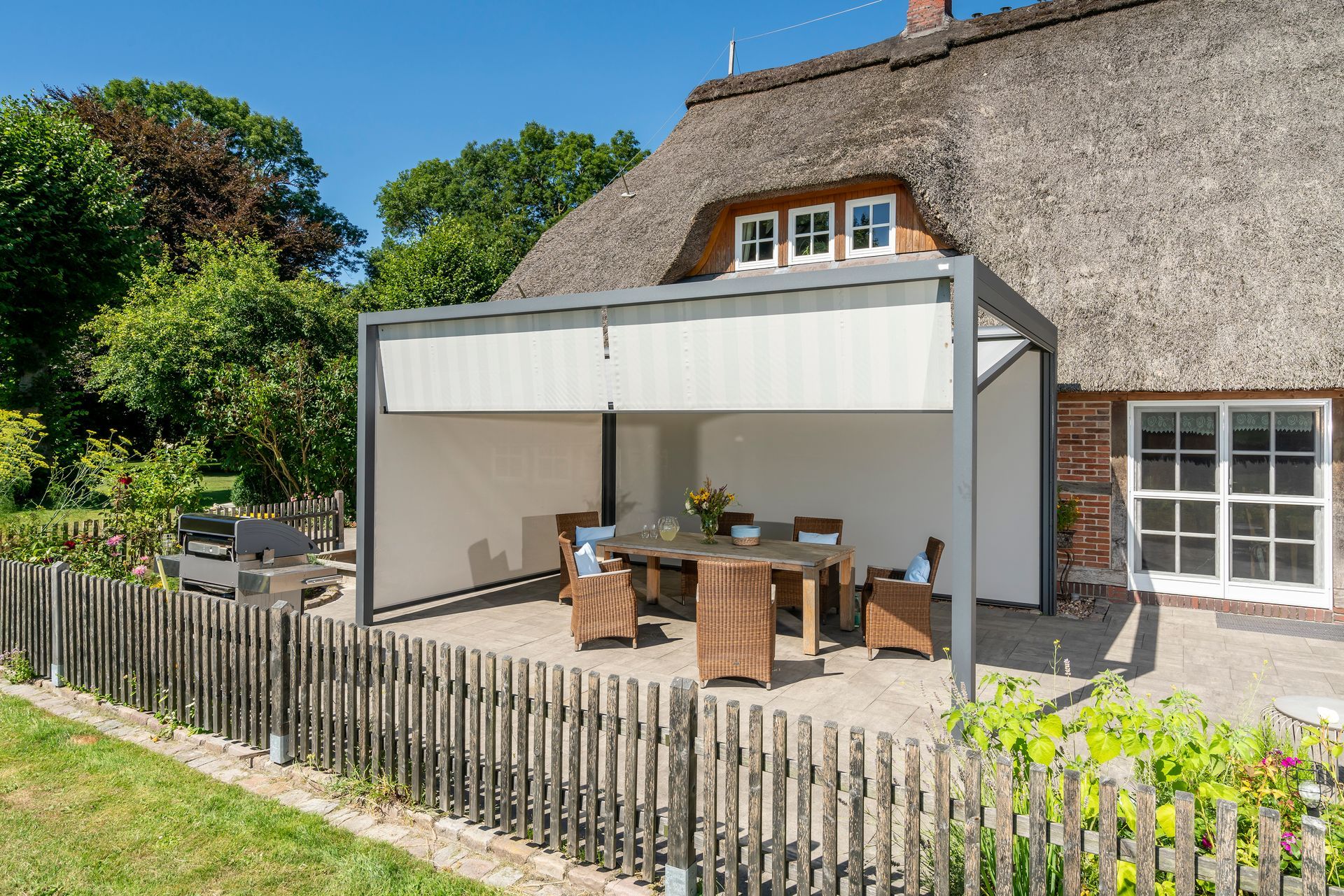 Ein Haus mit Reetdach und einer modernen, grau umrahmten Außenterrasse mit einem Esstisch auf einer Ziegelterrasse.
