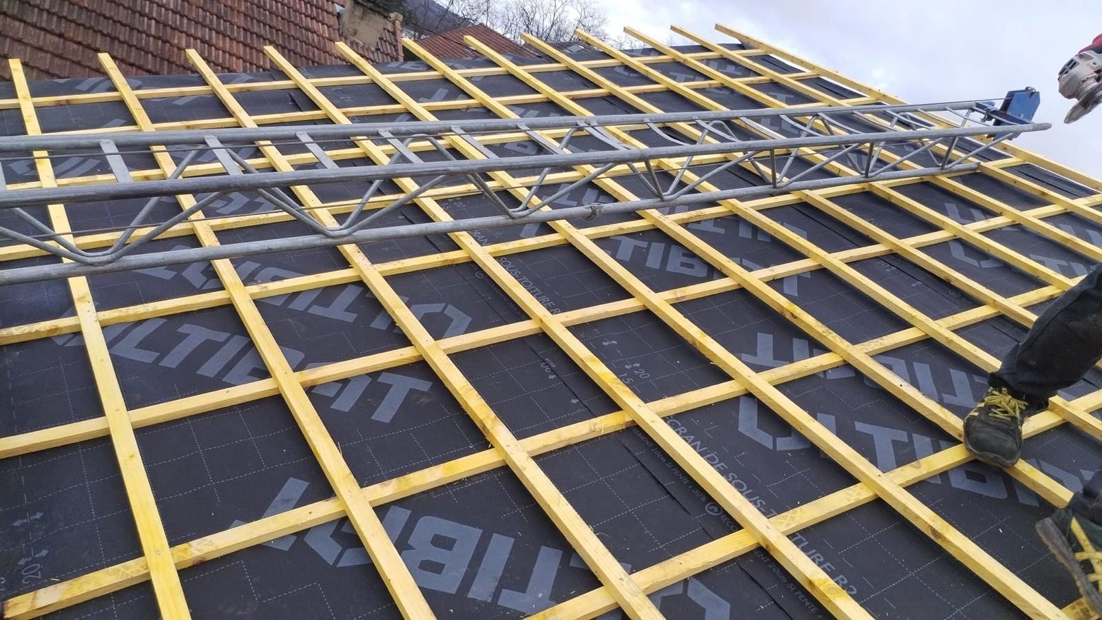 Toiture en construction : poutres en bois posées sur une sous-couche noire. Une échelle repose sur les poutres.