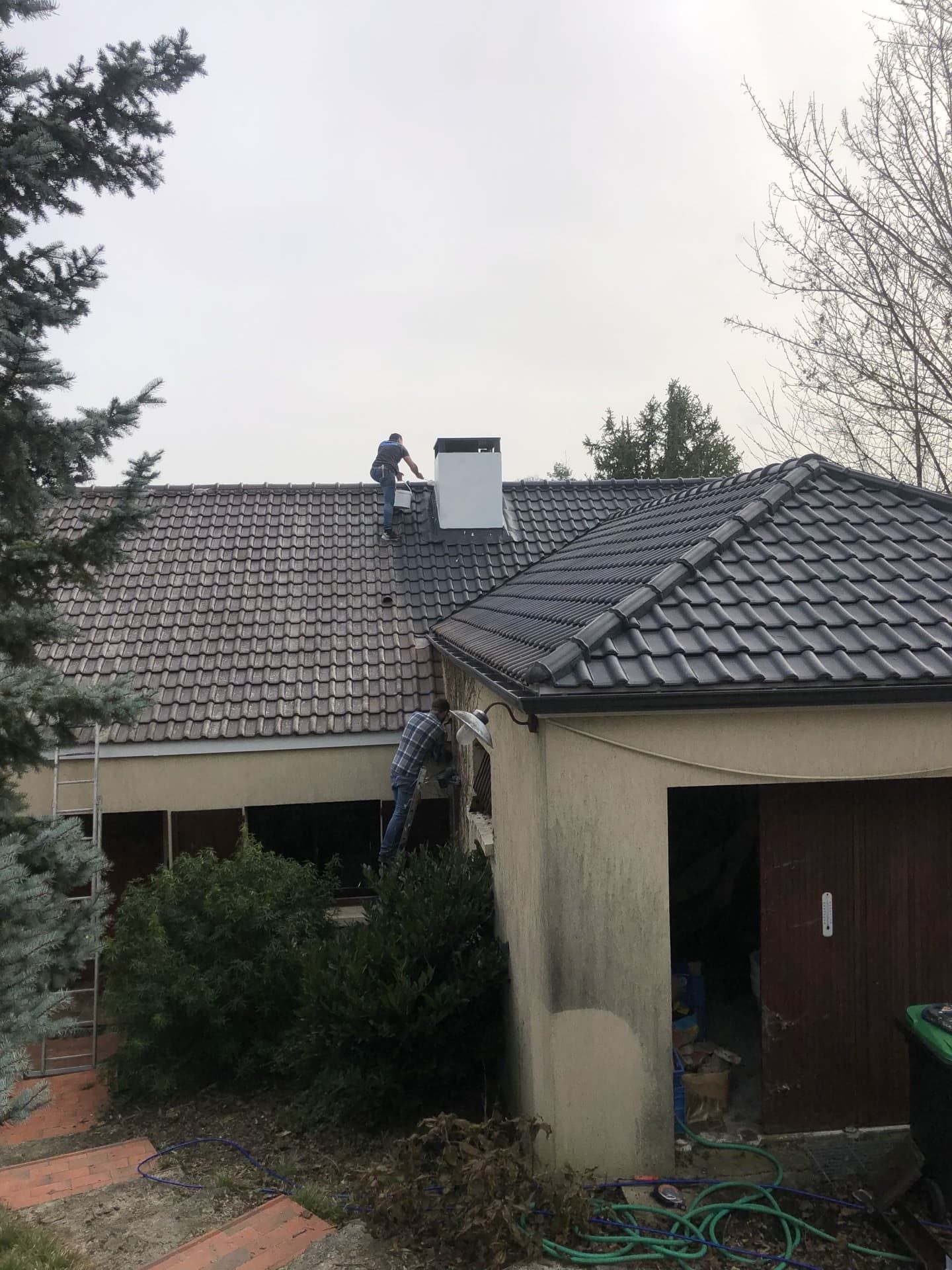 Deux ouvriers sur un toit, l'un près d'une cheminée, l'autre sur une échelle. Ciel couvert.