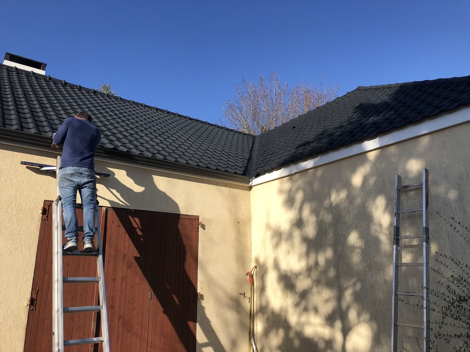 Une personne sur une échelle nettoie une gouttière sur un bâtiment au toit sombre.