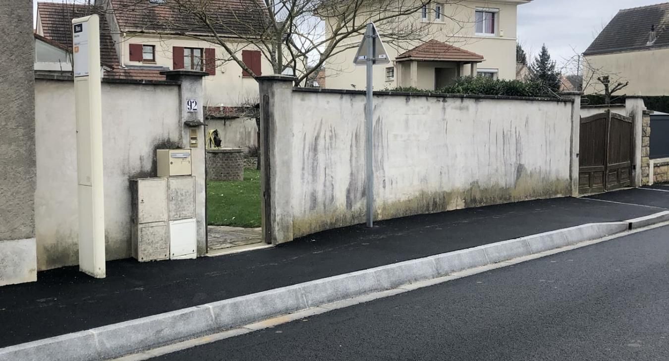 Un mur blanc borde une pelouse verte au bord d'une rue. Une boîte aux lettres est fixée au mur.