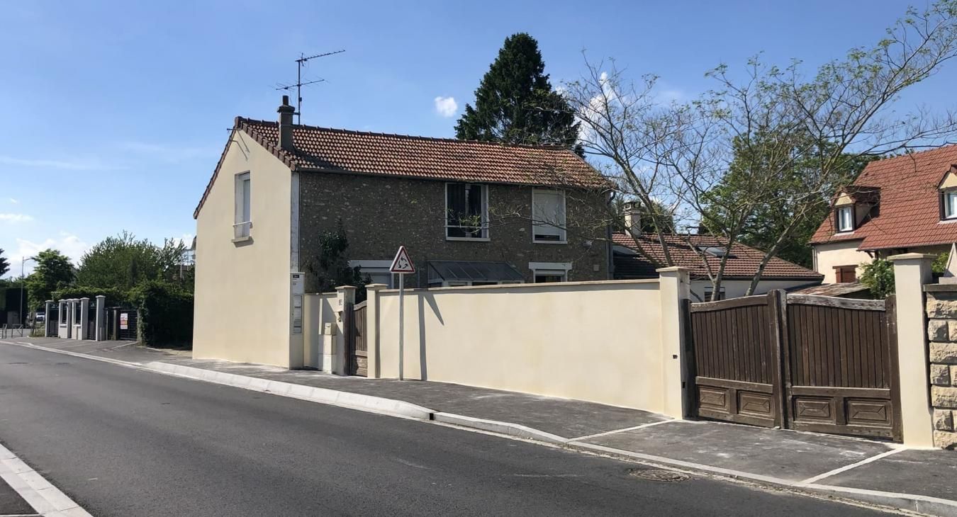Maison beige avec un toit de tuiles rouges derrière un mur et un portail beiges, le long d'une rue.