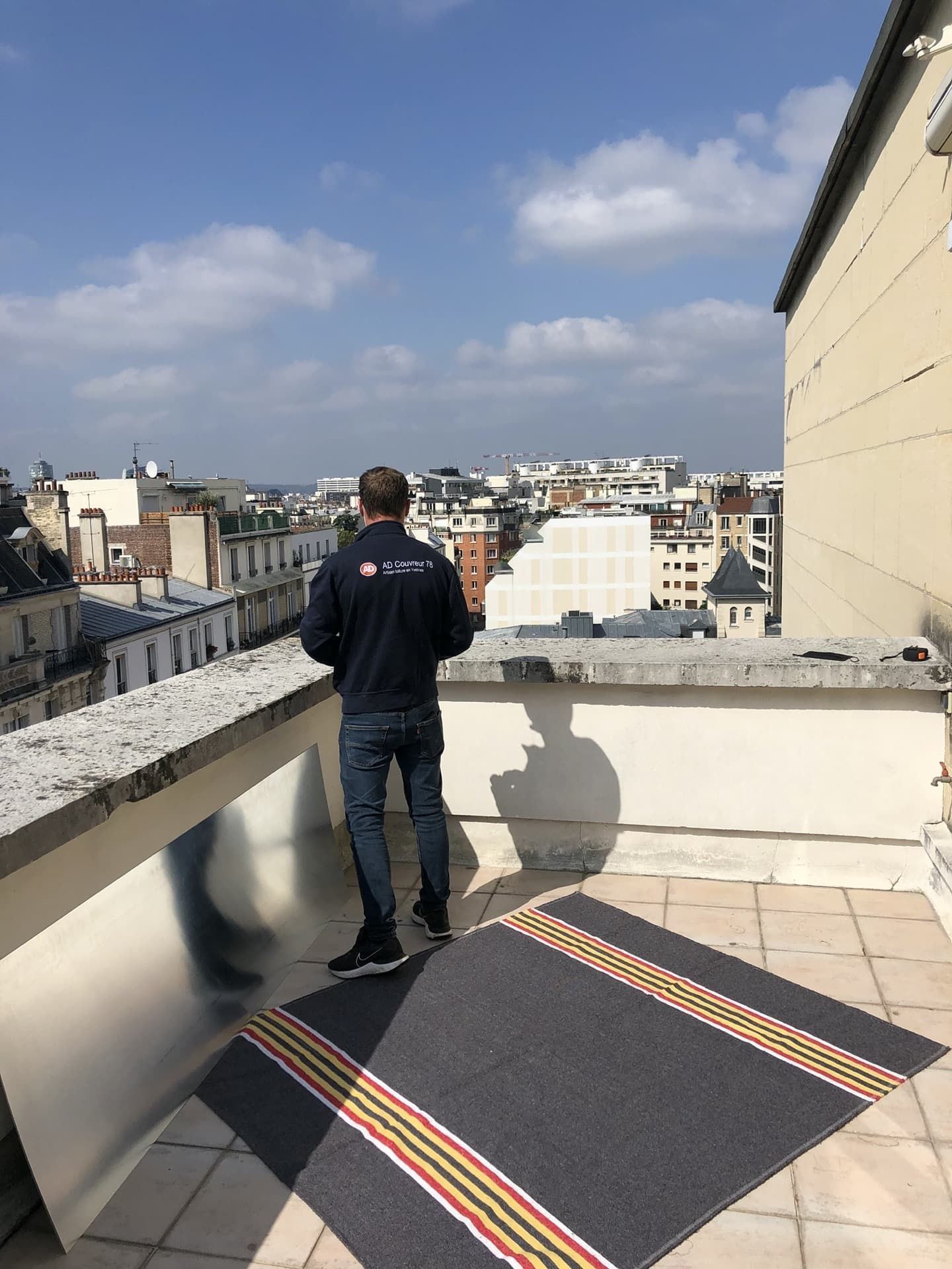 Un homme se tient sur un toit, contemplant la vue sur la ville. Tapis gris à rayures, journée ensoleillée.