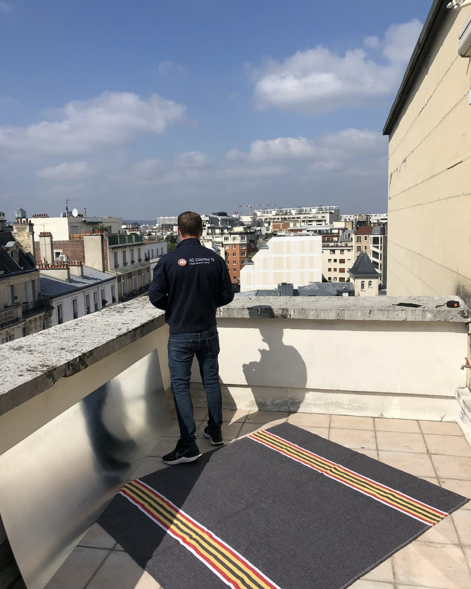 Un homme se tient sur un toit surplombant la ville, vêtu d'une veste bleu marine et d'un jean, près d'un tapis.