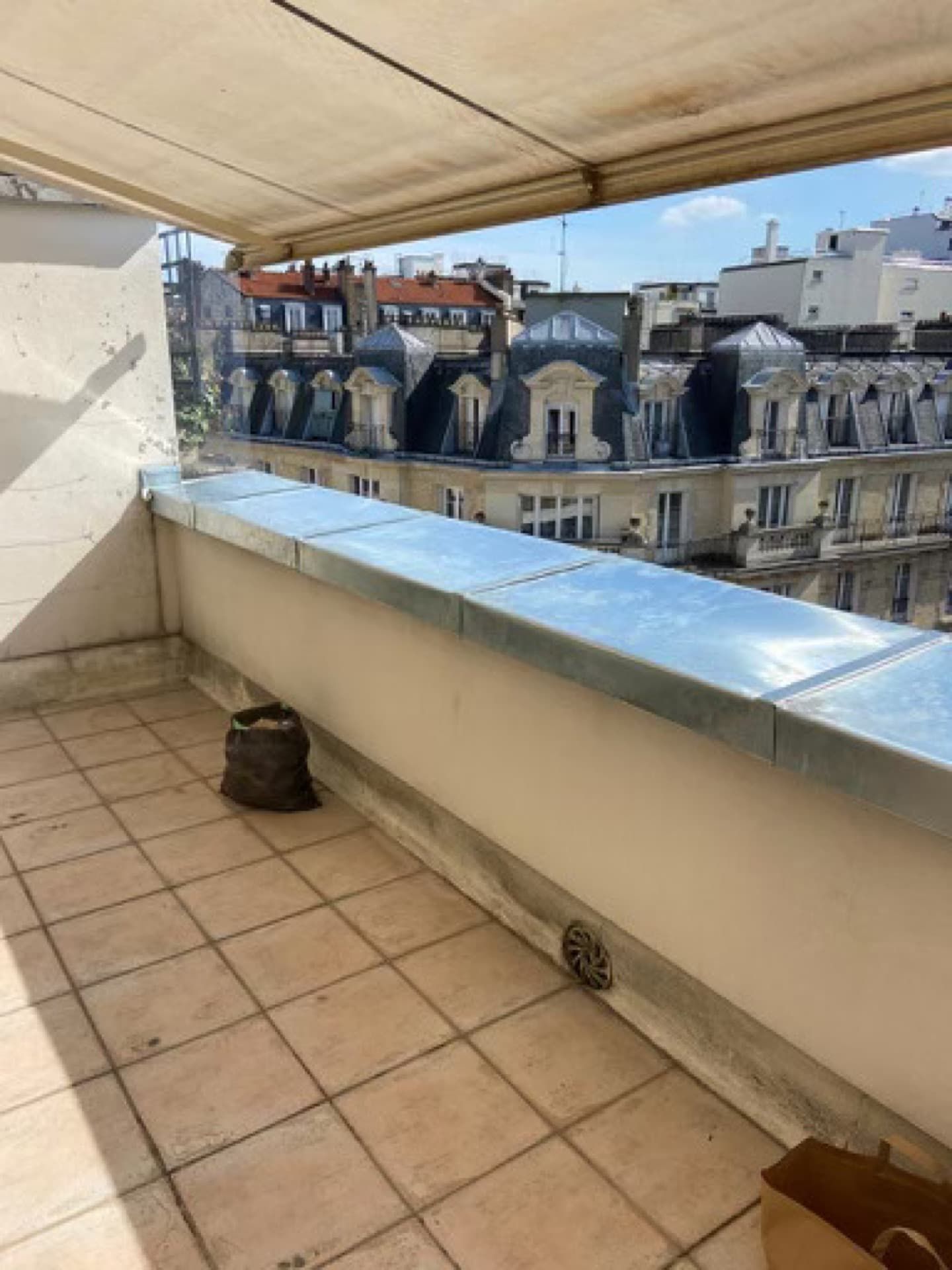 Balcon avec sol carrelé, bordure métallique et vue sur des immeubles aux toits rouges.