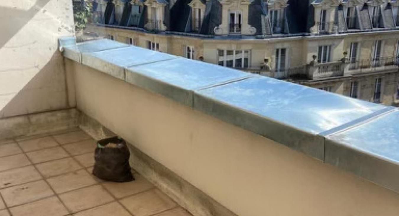 Un sac sombre posé sur un balcon carrelé avec une rambarde métallique, donnant sur un immeuble dans une ville européenne.