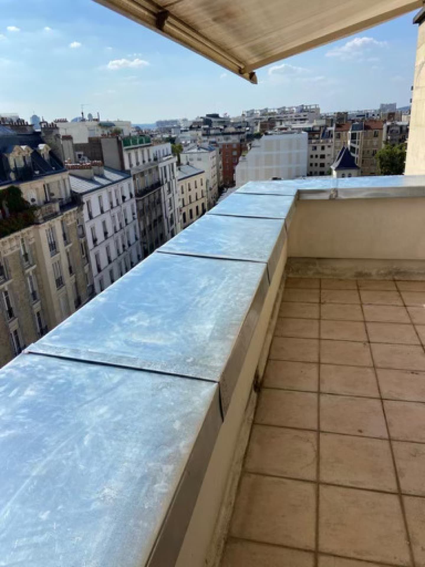 Balcon donnant sur les immeubles de la ville ; sol carrelé, rebord métallique gris, store rétractable.