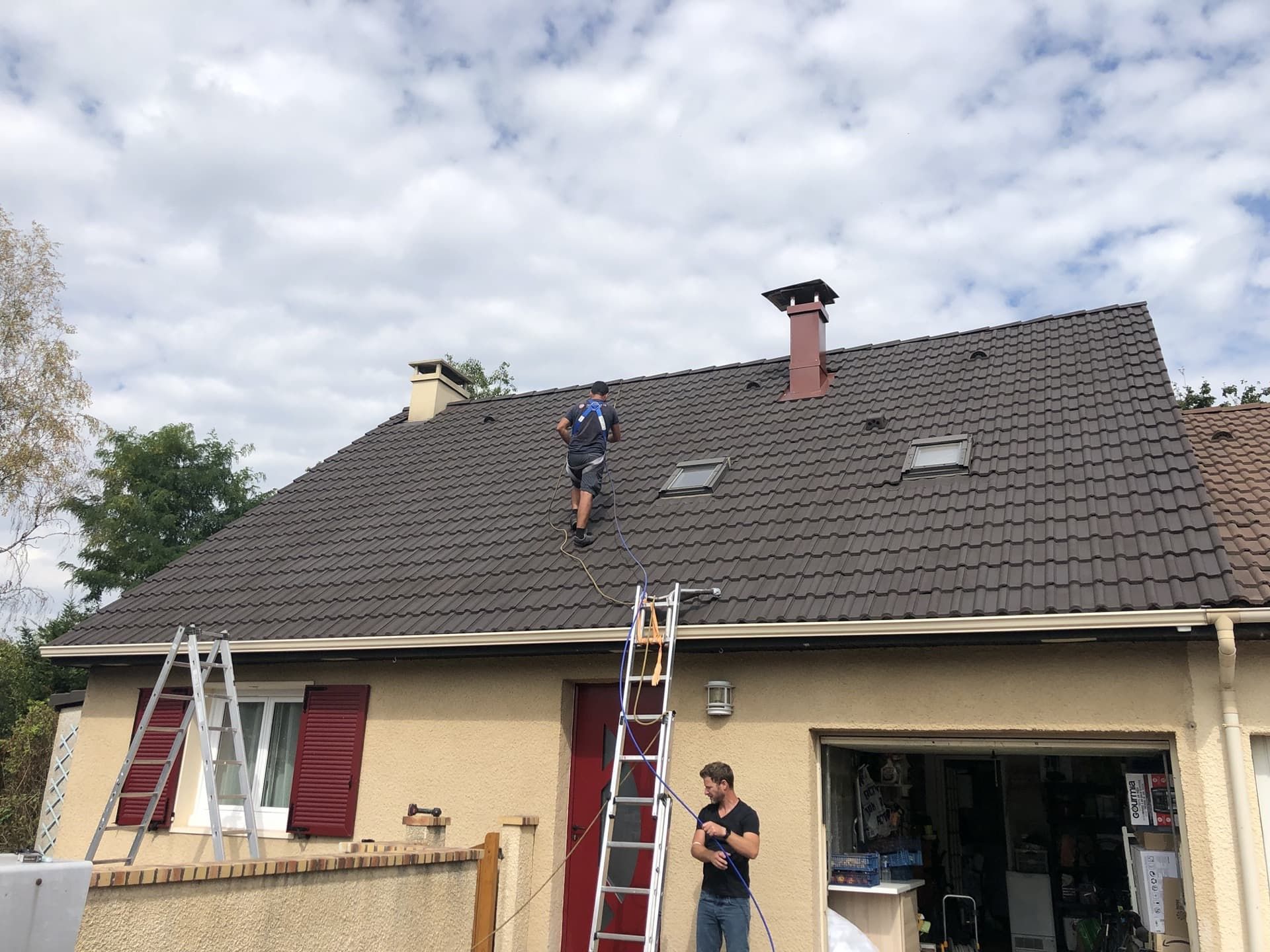 Deux ouvriers sur un toit de tuiles brunes, près des cheminées et des puits de lumière.