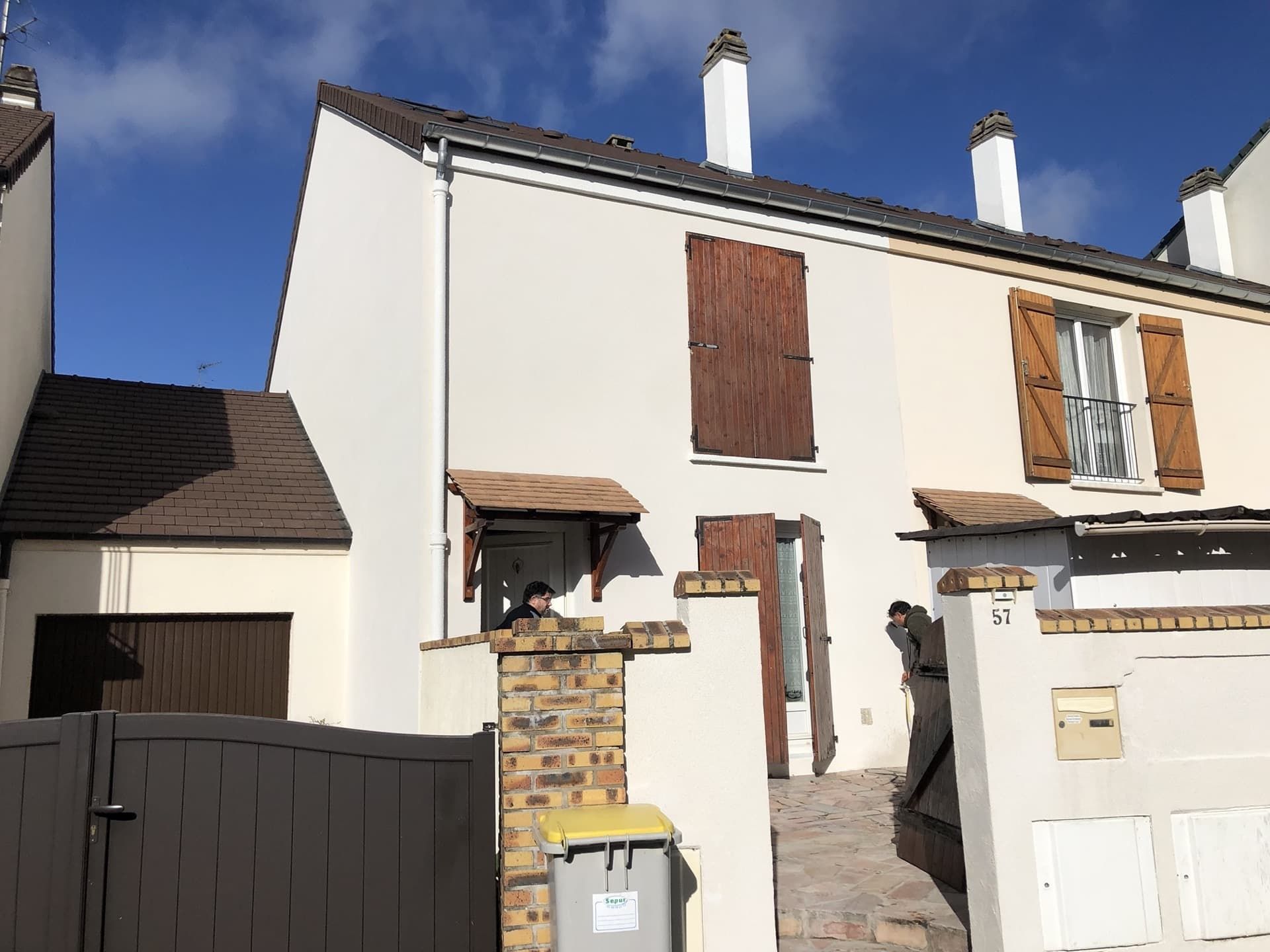Maison à deux étages avec volets bruns, toit brun et façade beige. Une petite colonne en briques soutient un auvent.