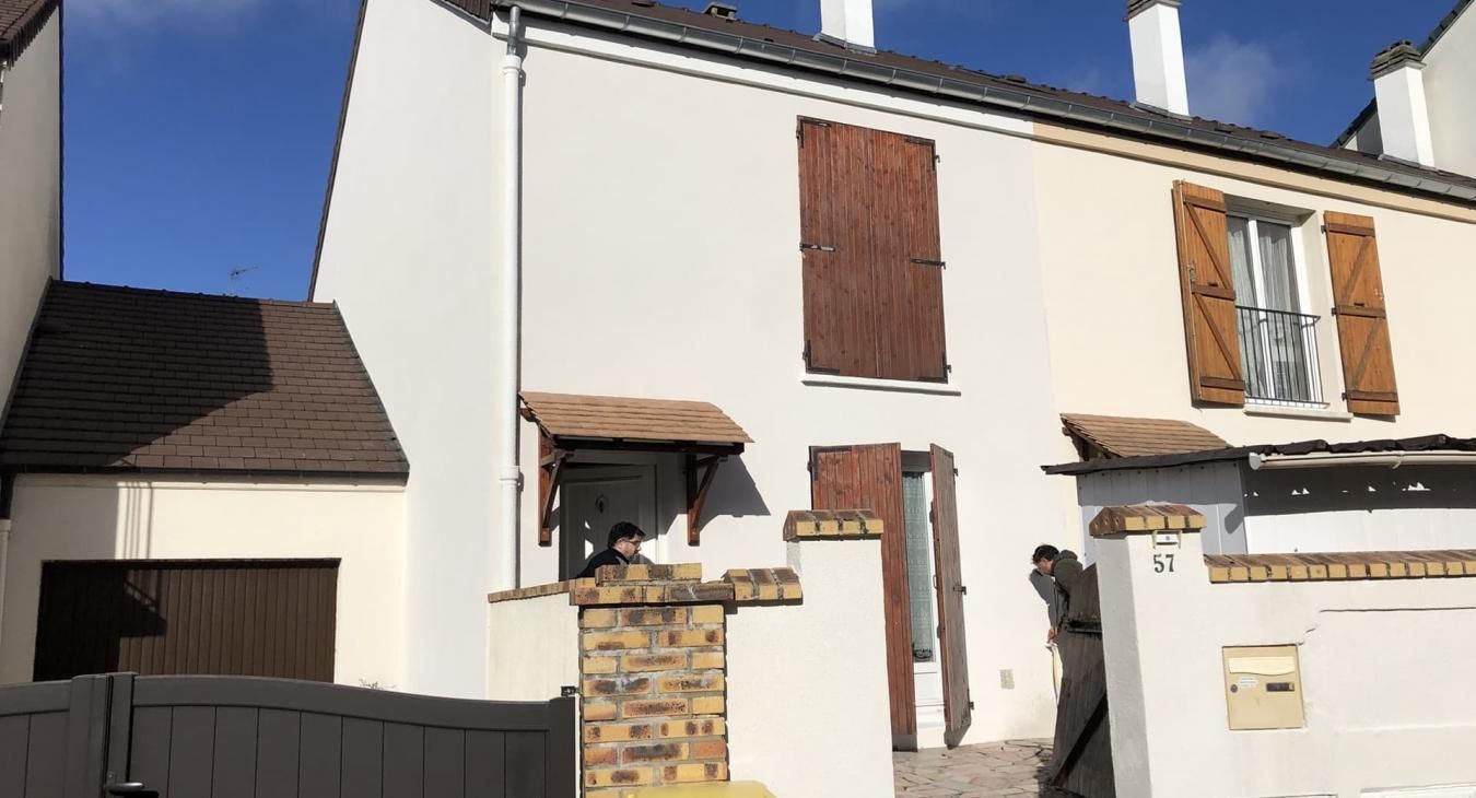 Maison mitoyenne aux murs couleur crème, volets marron, garage et petits auvents de véranda.