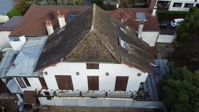 Vue aérienne d'une maison délabrée au toit de tuiles, aux murs blancs et aux volets bruns.