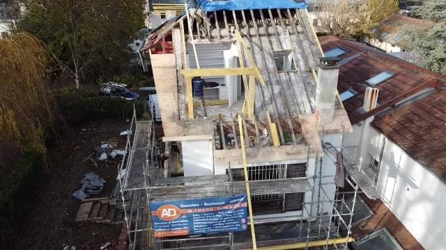 Maison en construction, toiture partiellement enlevée. Des échafaudages entourent le bâtiment, un ouvrier est visible.
