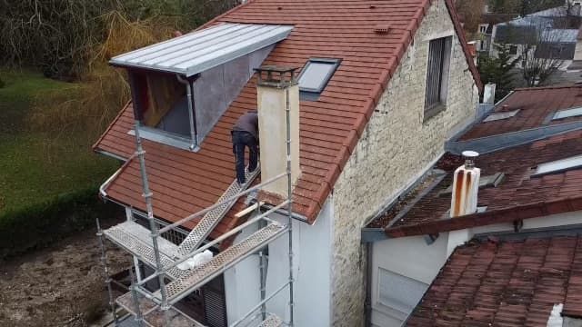 Une personne travaille sur un toit équipé d'échafaudages, près d'une cheminée et d'un puits de lumière.