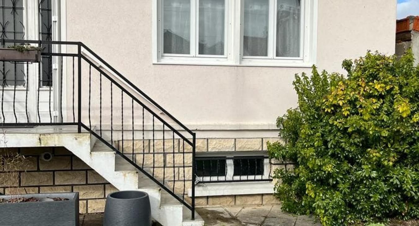 Extérieur d'un bâtiment avec escalier, rambarde noire et fenêtre. Un buisson vert se trouve à droite.