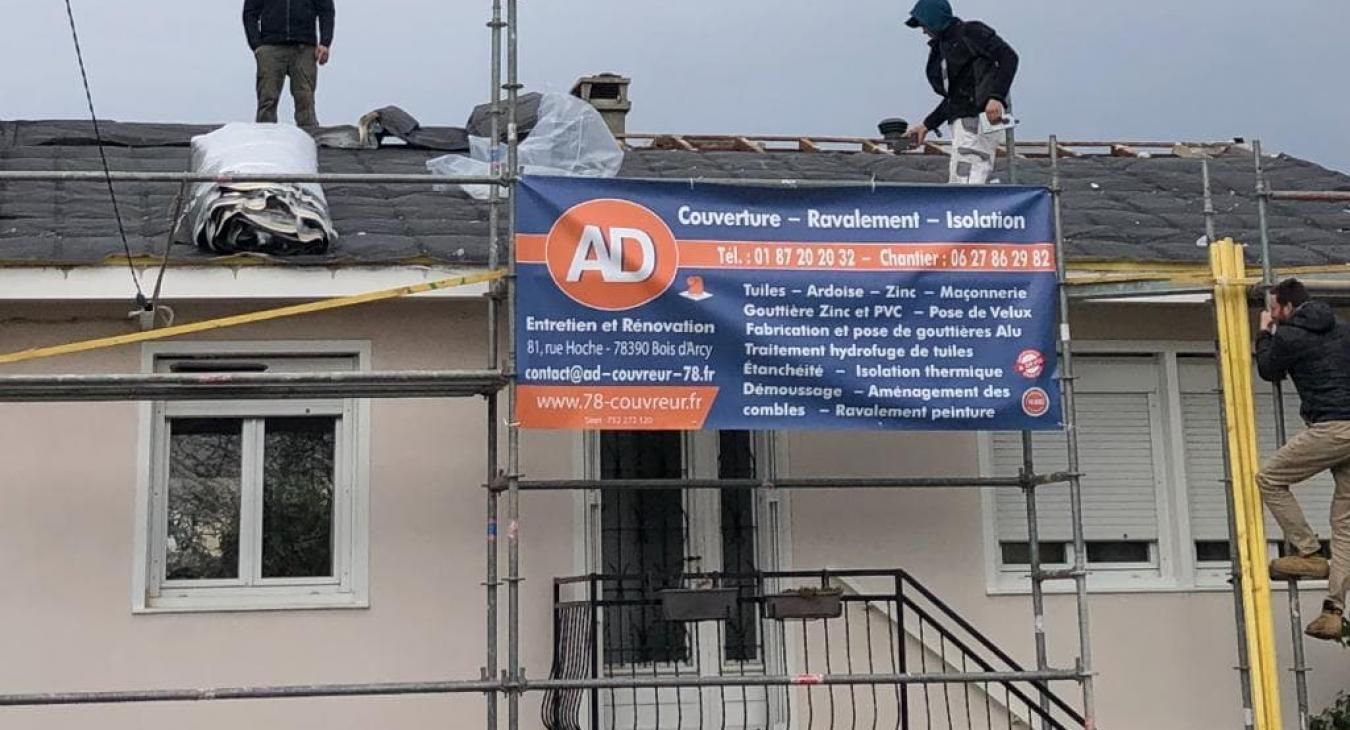 Des ouvriers du bâtiment réparent des tuiles sur un toit. Un échafaudage soutient une banderole pour AD Couverture.