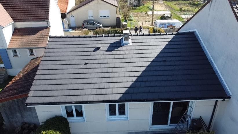 Vue d'un toit de tuiles gris foncé sur une maison aux murs, fenêtres et cheminée blancs.
