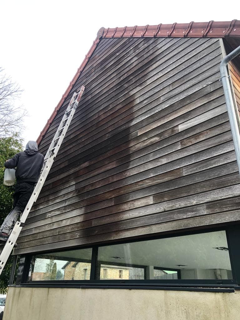 Une personne sur une échelle lave le revêtement en bois d'une maison. La zone assombrie indique le nettoyage.