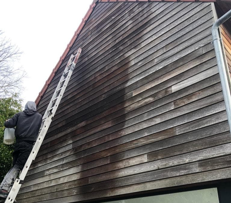 Une personne perchée sur une échelle lave à l'aide d'un seau la façade en bois patinée d'une maison.