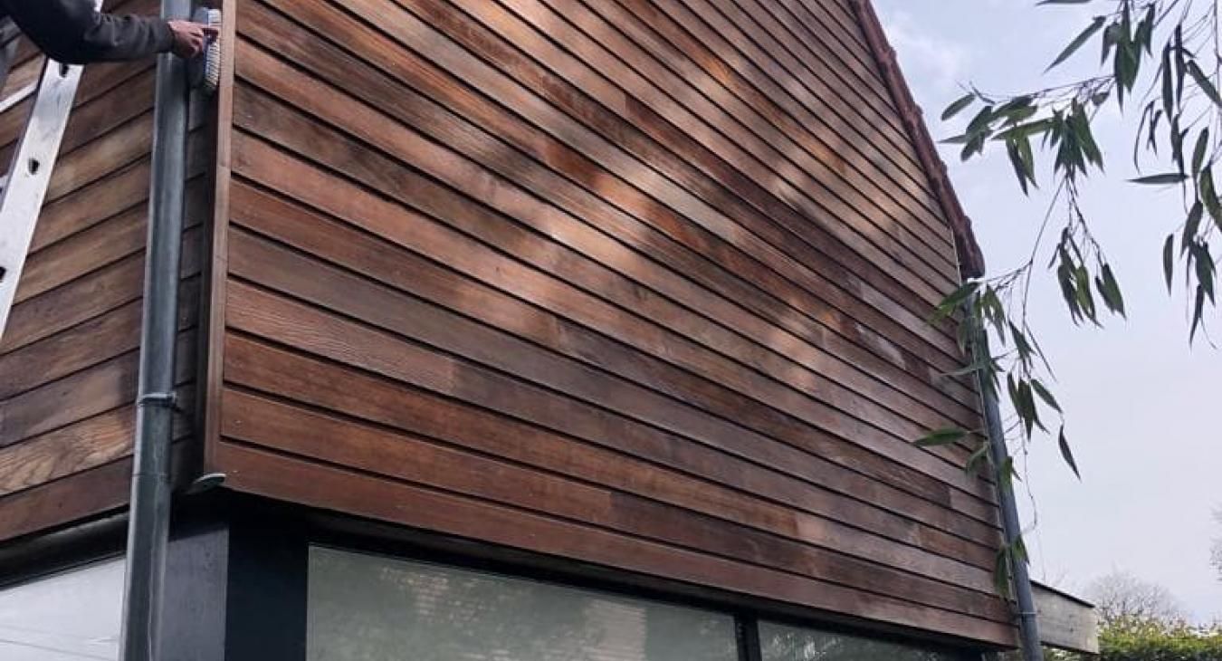 Extérieur d'une maison à panneaux de bois avec une échelle, une gouttière et une fenêtre, arbres en arrière-plan.