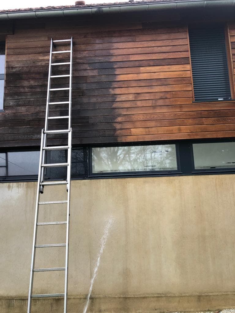 Une personne nettoie à haute pression la moitié supérieure du bardage d'un bâtiment en bois.