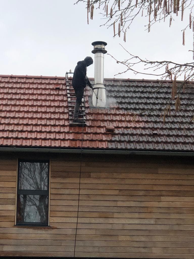 Une personne ramonait une cheminée sur un toit de tuiles rouges, debout sur une échelle. Bardage en bois en dessous.