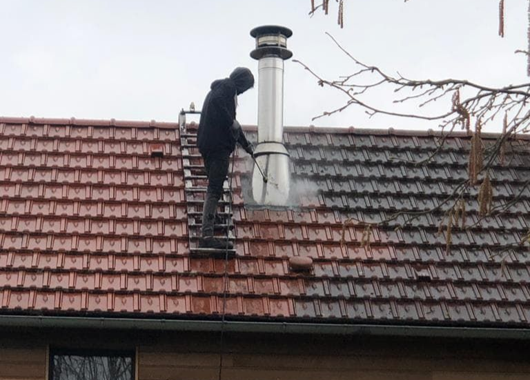 Une personne sur une échelle nettoie au nettoyeur à haute pression un toit de tuiles rouges près d'une cheminée.
