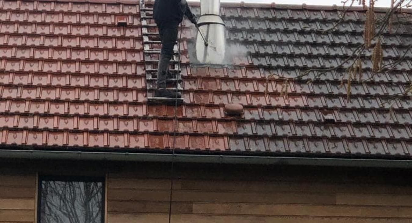 Une personne sur une échelle nettoie au jet d'eau un toit de tuiles rouges, enlevant les algues et la saleté.