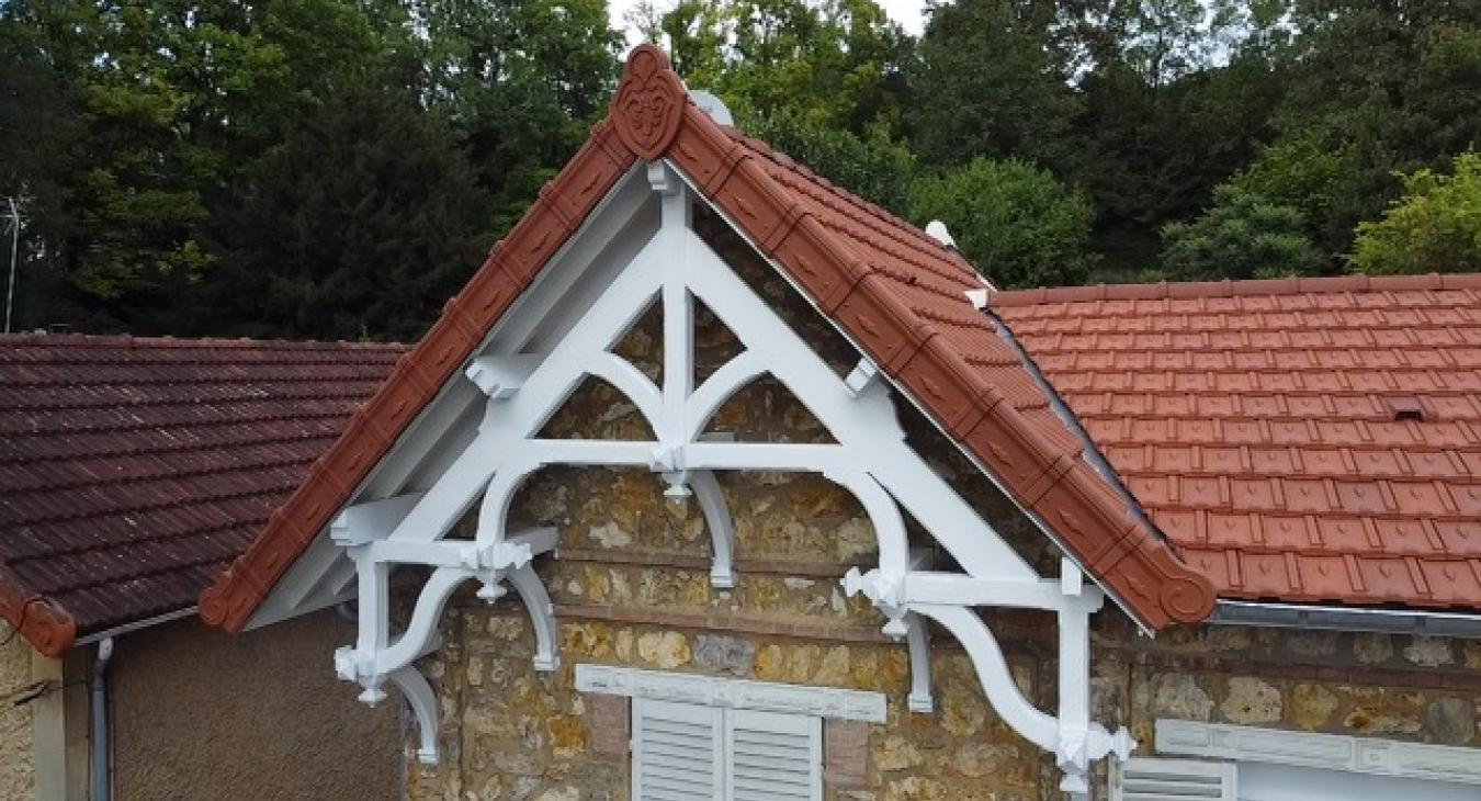 Pignon décoratif en bois blanc au-dessus d'un bâtiment en pierre au toit de tuiles rouges, avec une forêt en arrière-plan.