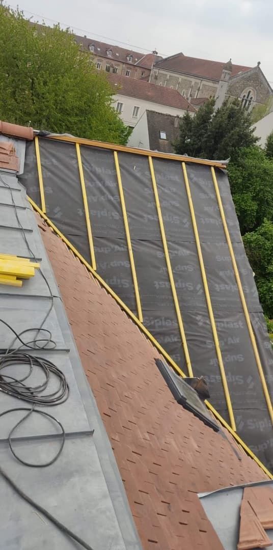 Toiture en construction avec sous-couche noire, poutres en bois et tuiles rouges. Un bâtiment est visible en arrière-plan.
