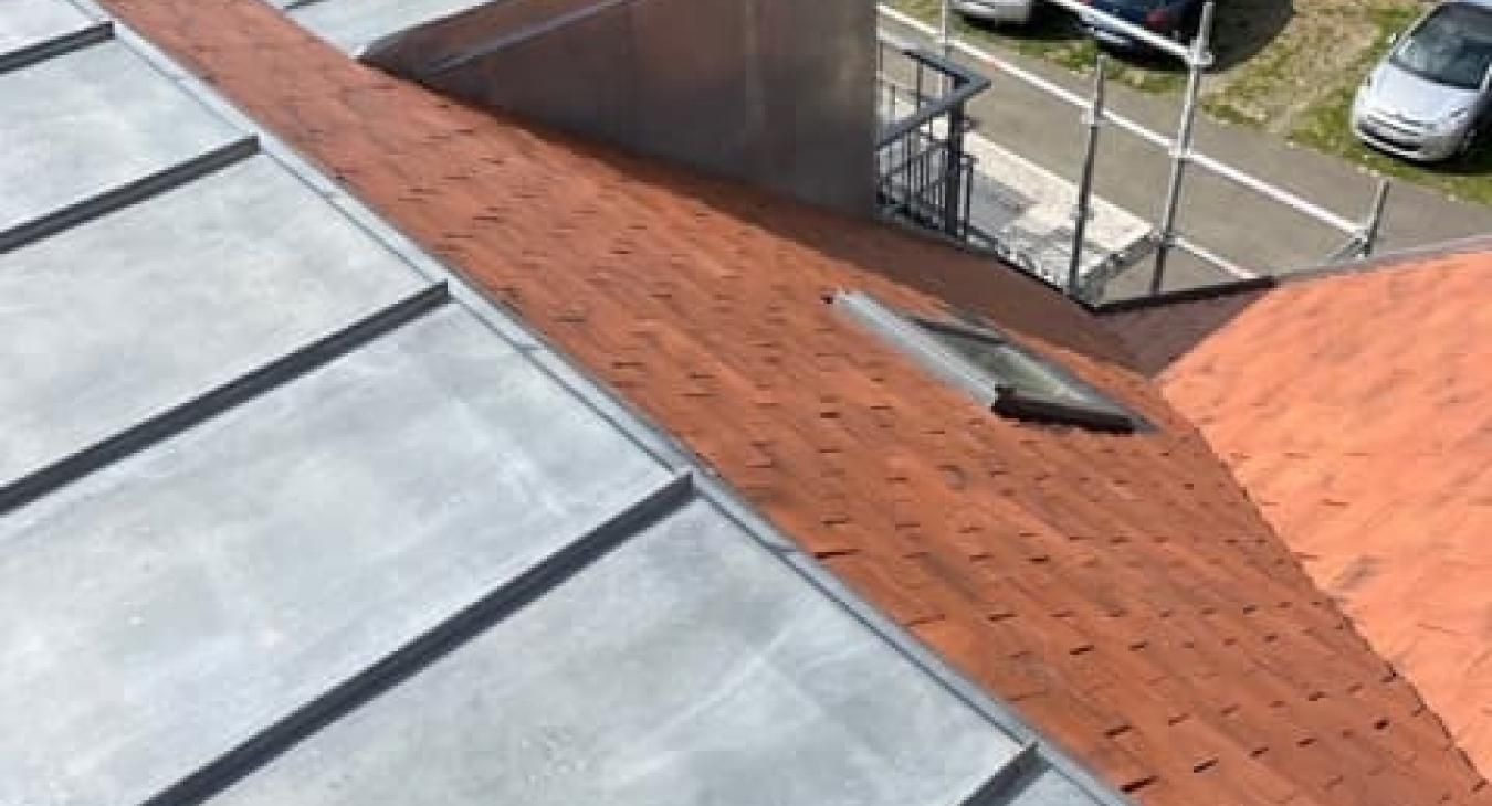 Vue aérienne d'un toit recouvert de tuiles en terre cuite rouge et de panneaux métalliques argentés. On aperçoit une lucarne.