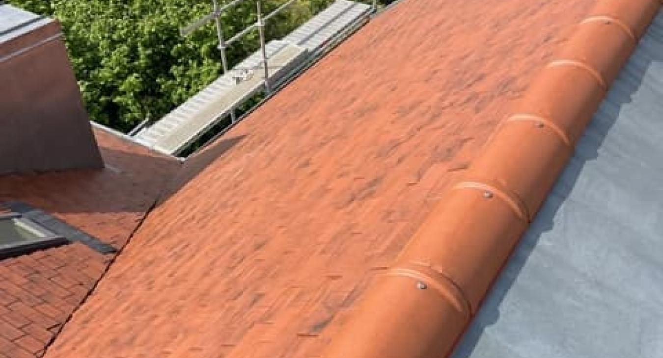 Toiture en tuiles rouges avec cheminée et solins métalliques gris. Des échafaudages sont visibles en arrière-plan.