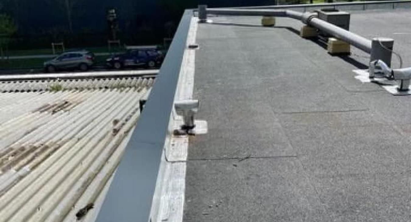 Un toit plat et noir avec une rambarde métallique. À côté, un toit en tôle ondulée. Une voiture est visible dans la rue.