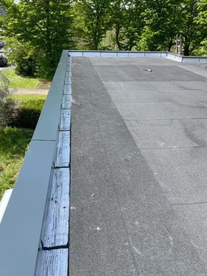 Toit plat recouvert d'une toiture sombre, bordé de gris et orné de boiseries. Arbres en arrière-plan.