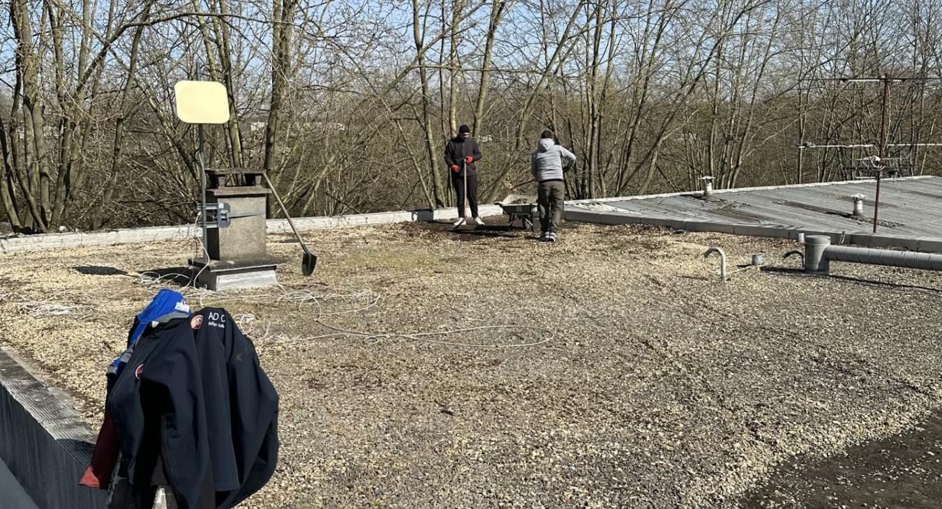 Trois personnes travaillent sur un toit de gravier près des arbres. Une veste bleue est posée sur le bord.