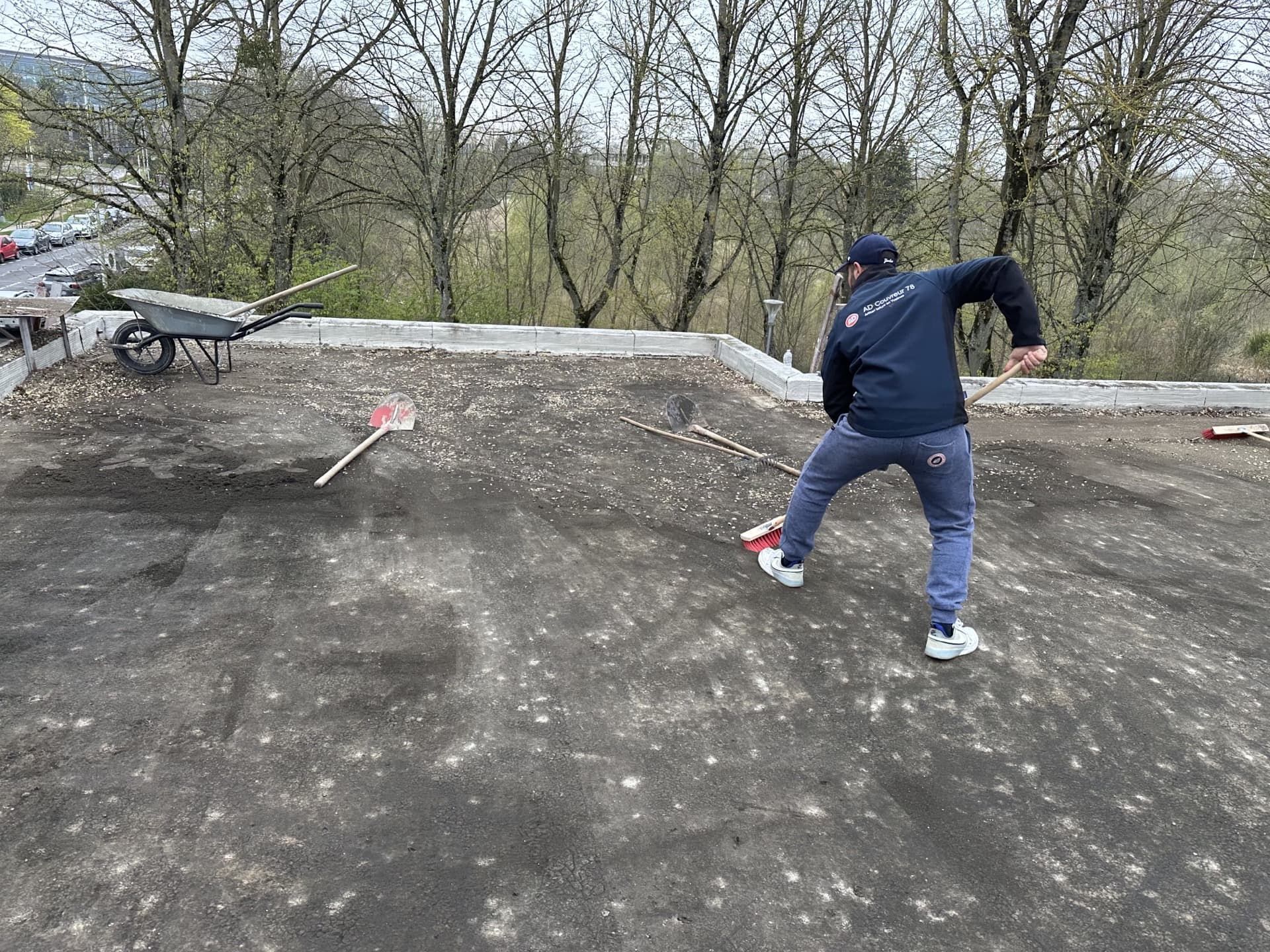 Personne balayant des débris sur un toit plat. Surface grise, arbres en arrière-plan, chemise bleue, pantalon foncé.