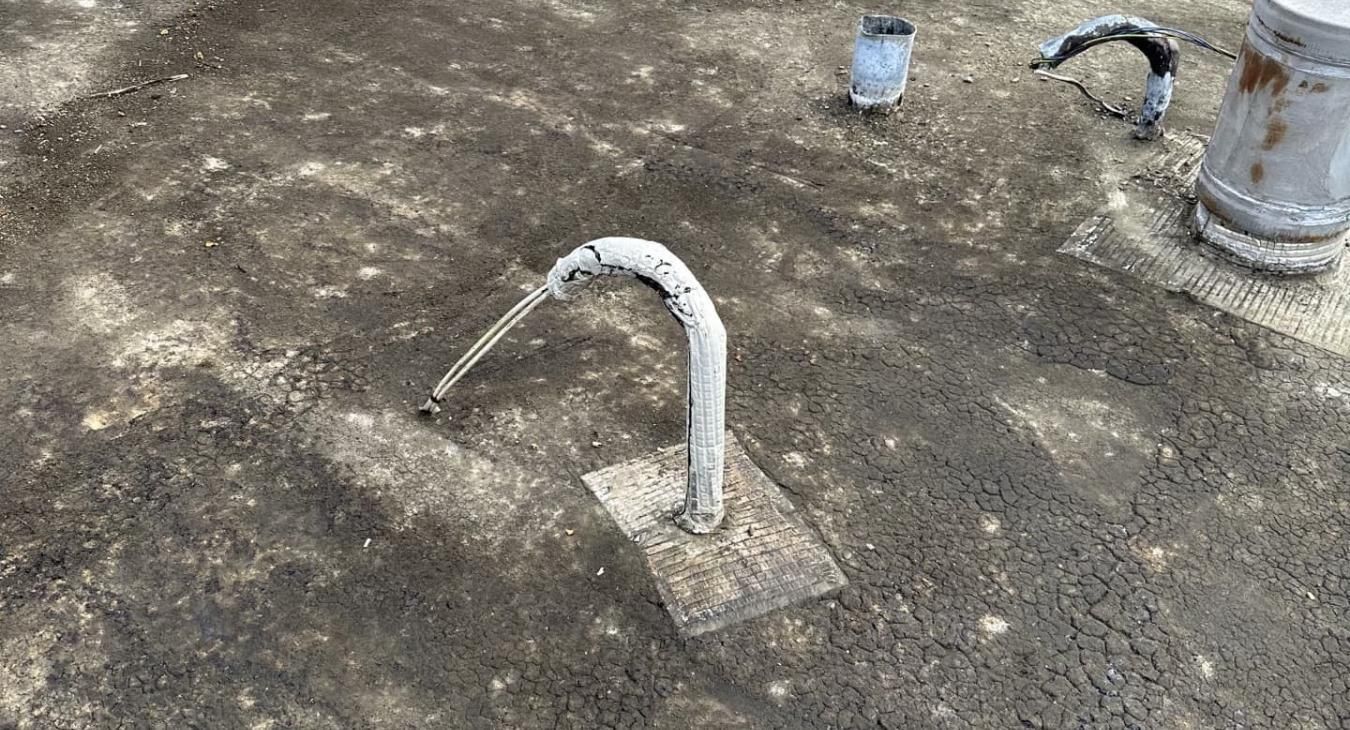 Un tuyau métallique courbé émerge du sol sur une surface sombre et texturée, avec d'autres objets métalliques à proximité.