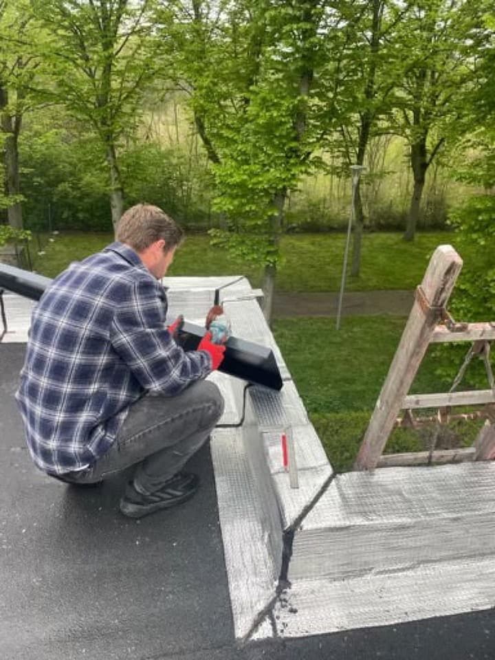 Une personne sur un toit utilise une meuleuse sur un tuyau noir, des arbres sont visibles en arrière-plan.