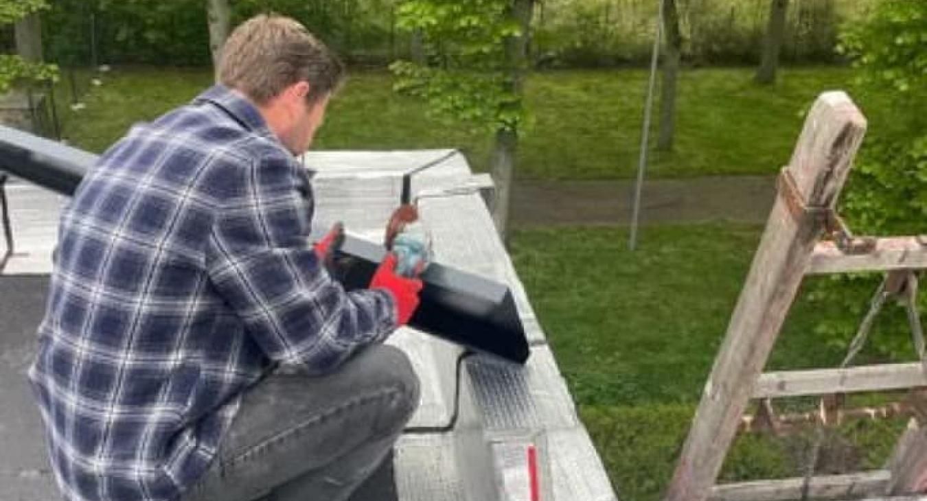 Un homme en chemise à carreaux se trouve sur un toit, réparant une gouttière noire.