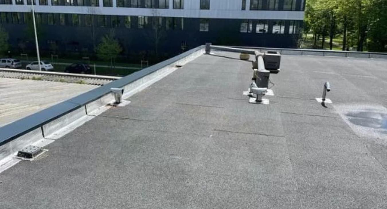 Toit plat avec bordure métallique, aérations et un bâtiment en arrière-plan. Gris et noir.