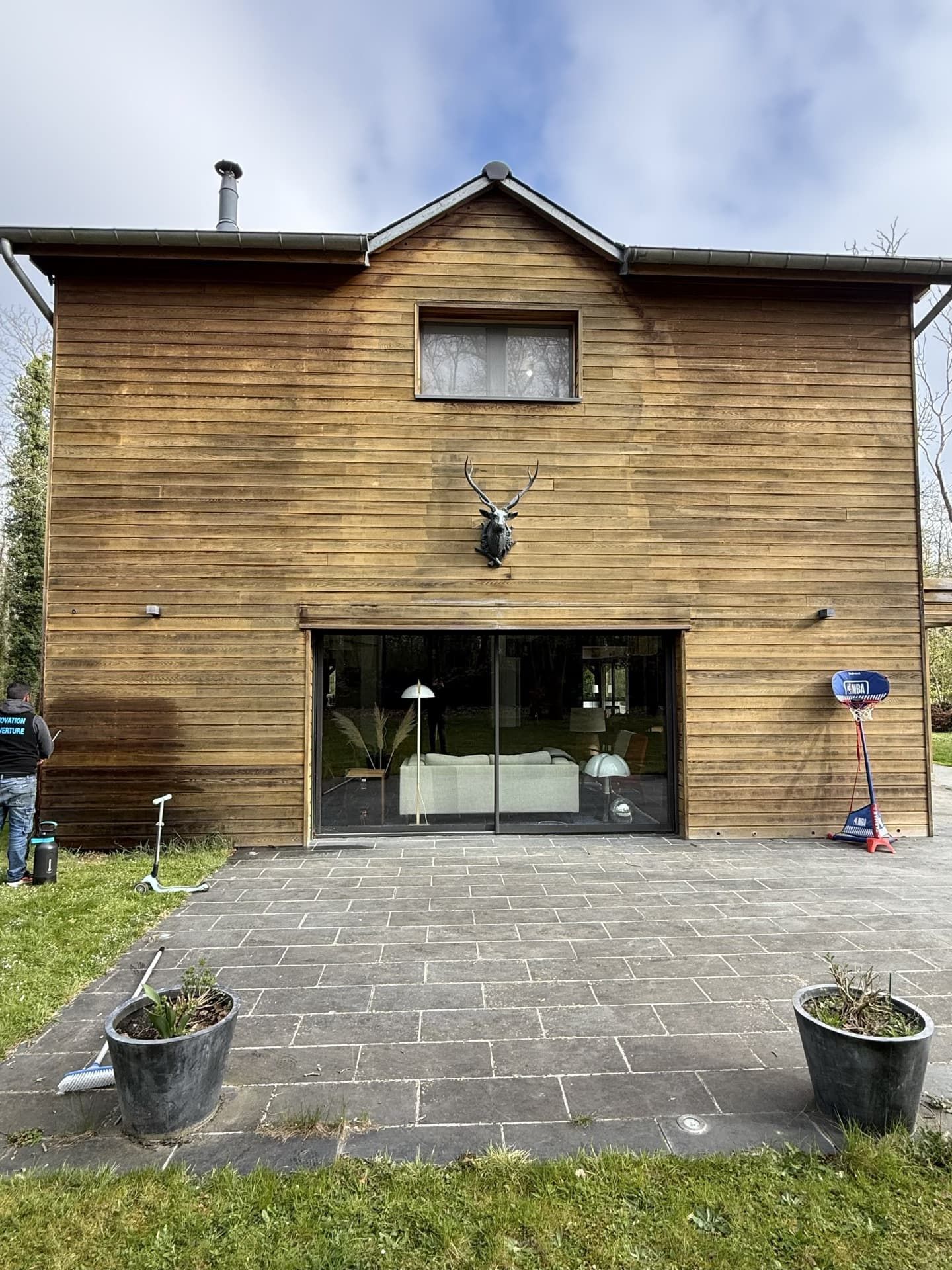 Maison en bois avec de grandes portes vitrées. Personne à gauche. Panier de basket à droite. Plantes en pot au premier plan.