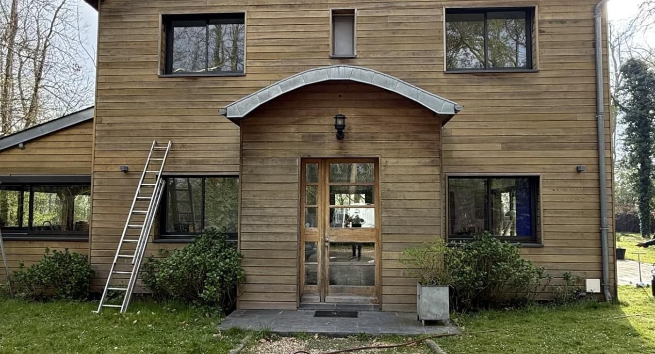 Maison en bois aux fenêtres sombres, entrée cintrée, porte, petites plantes et une échelle appuyée contre le mur.