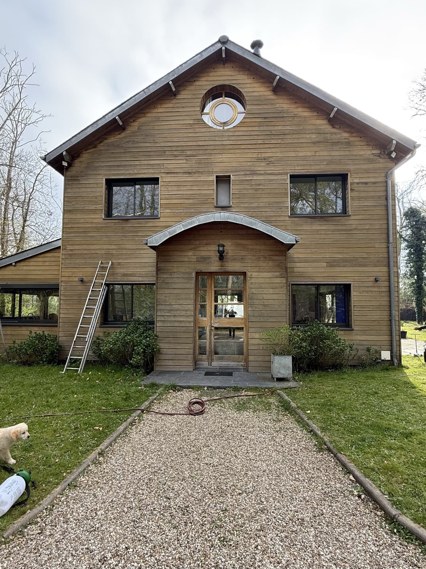 Maison en bois avec chemin de gravier, échelle et végétation. Extérieur en bois clair.
