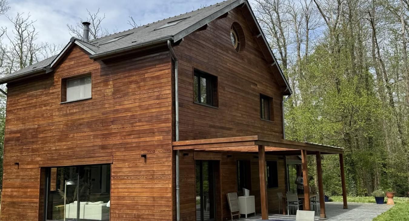 Maison en bois de deux étages avec véranda et arbres environnants.