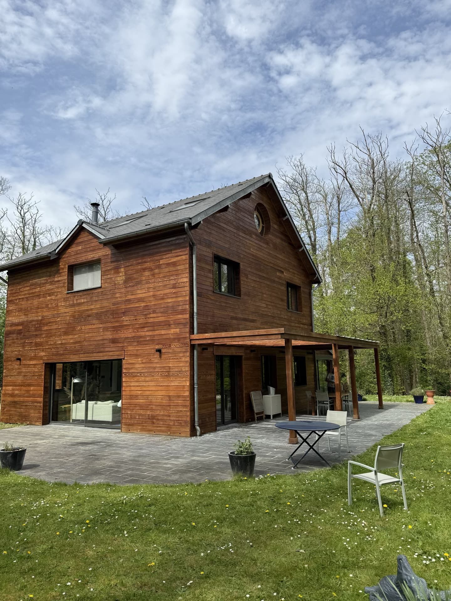 Maison en bois de deux étages avec toit sombre et patio, située sur une pelouse verdoyante arborée.