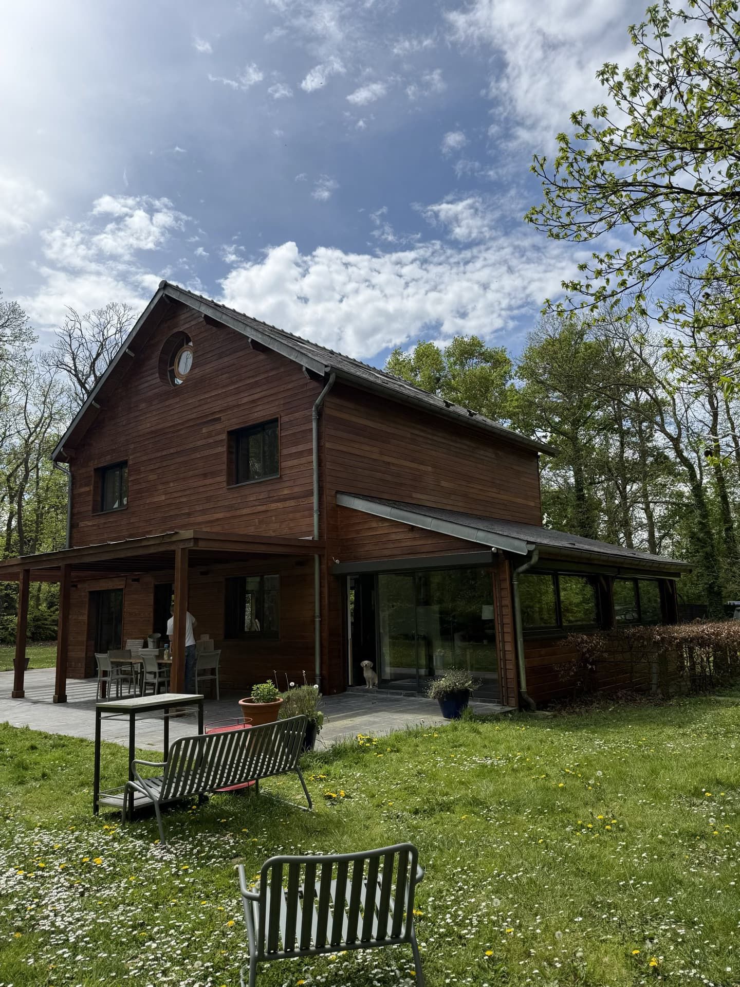 Maison en bois de deux étages avec patio couvert, située dans un jardin gazonné sous un ciel partiellement nuageux.