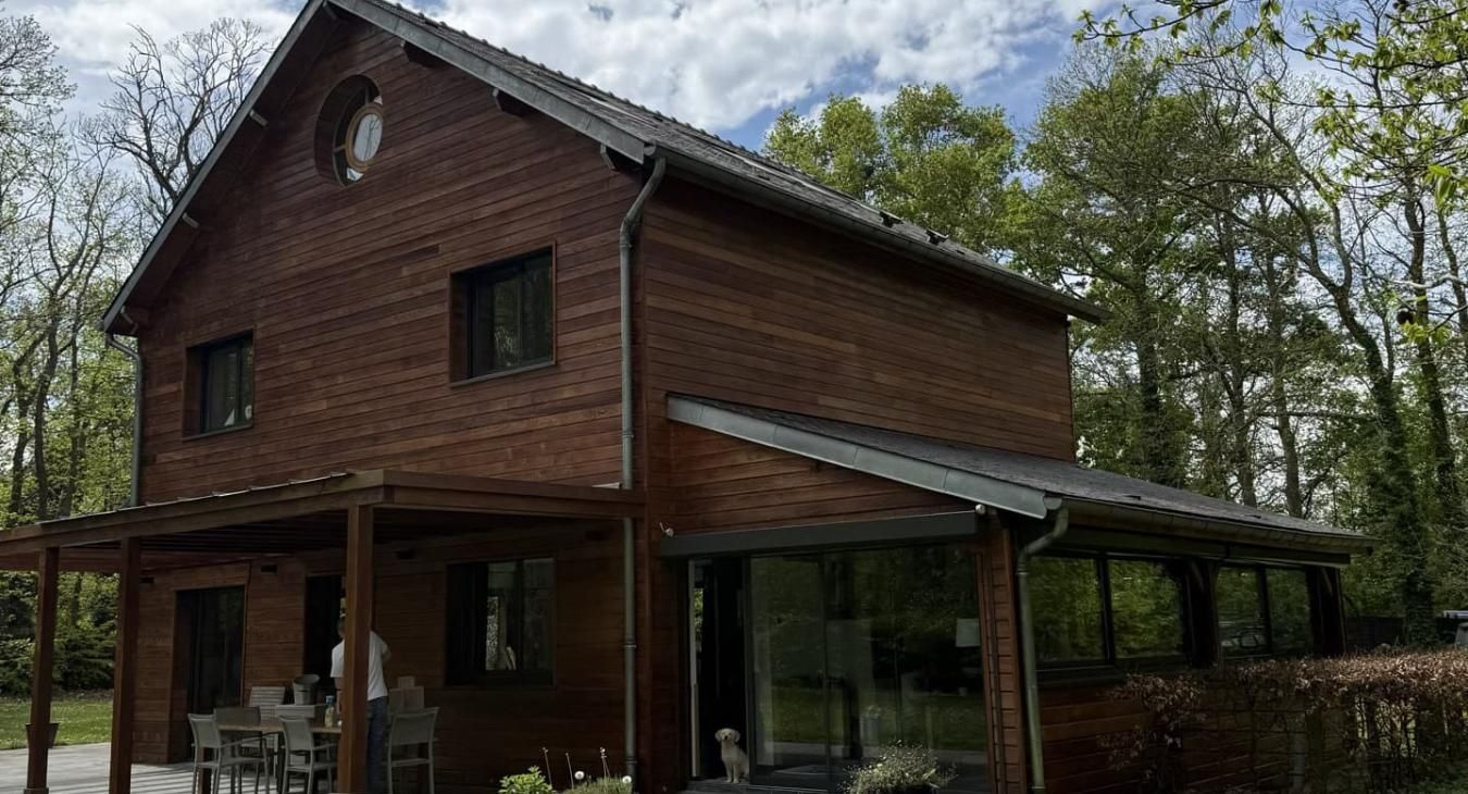 Maison en bois à deux étages avec un porche couvert, entourée d'arbres sous un ciel nuageux.